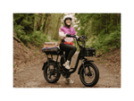 A woman sitting on a black RadRunner Plus electric cargo utility bike wearing a white helmet, pink jacket and grey pants.