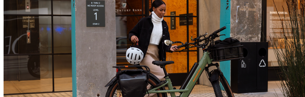 A woman walks out of an office building holding a helmet toward her green electric bike.