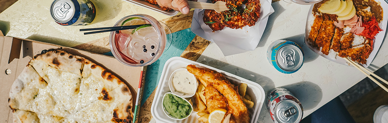 Various takeout dishes and sodas arrayed on a table, including pizza and fish and chips.