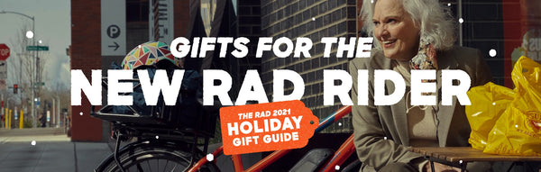 An older woman admires her RadMission outside a cafe. Banner text reads "Gift fo the New Rad Rider" and "Rad Power Bikes Holiday Gift Guide"