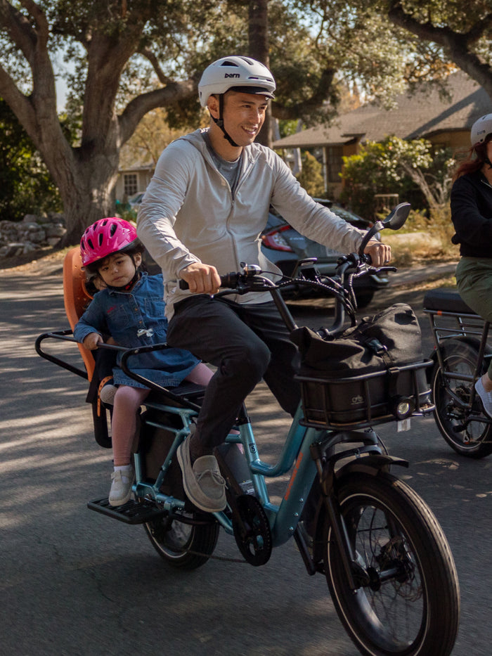 Man riding a metallic blue RadWagon 5 electric cargo bike with two small children riding on the back with passenger accessories installed. 