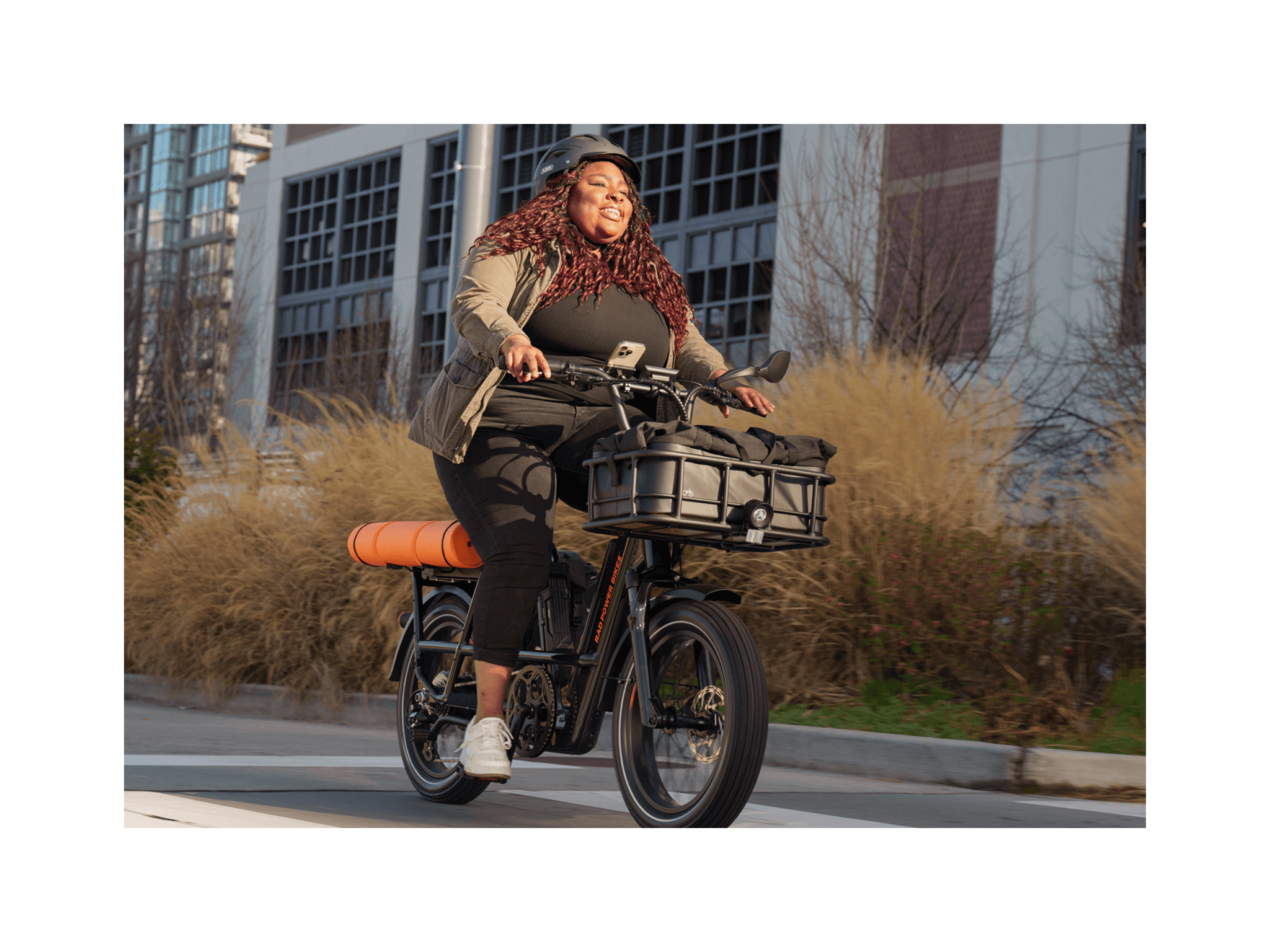 A woman wearing a black outfit with green jacket and a black bike helmet riding a green electric bike on a road in a city setting. 