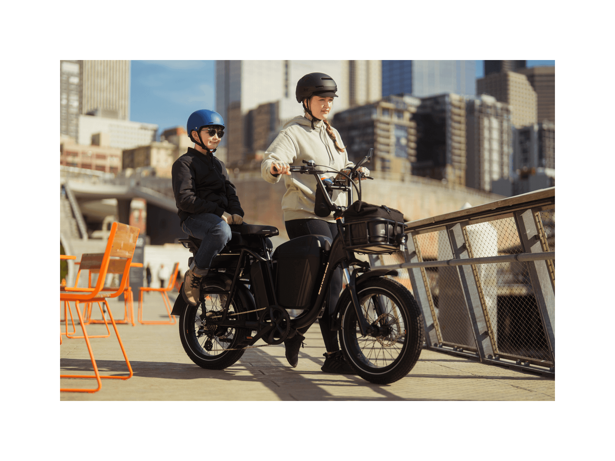 A woman wearing a tan hoodie and black helmet holding the handlebars of a matte black electric bike with a boy wearing a black shirt and jeans and blue helmet sitting on the back. On a bridge in an urban setting. 