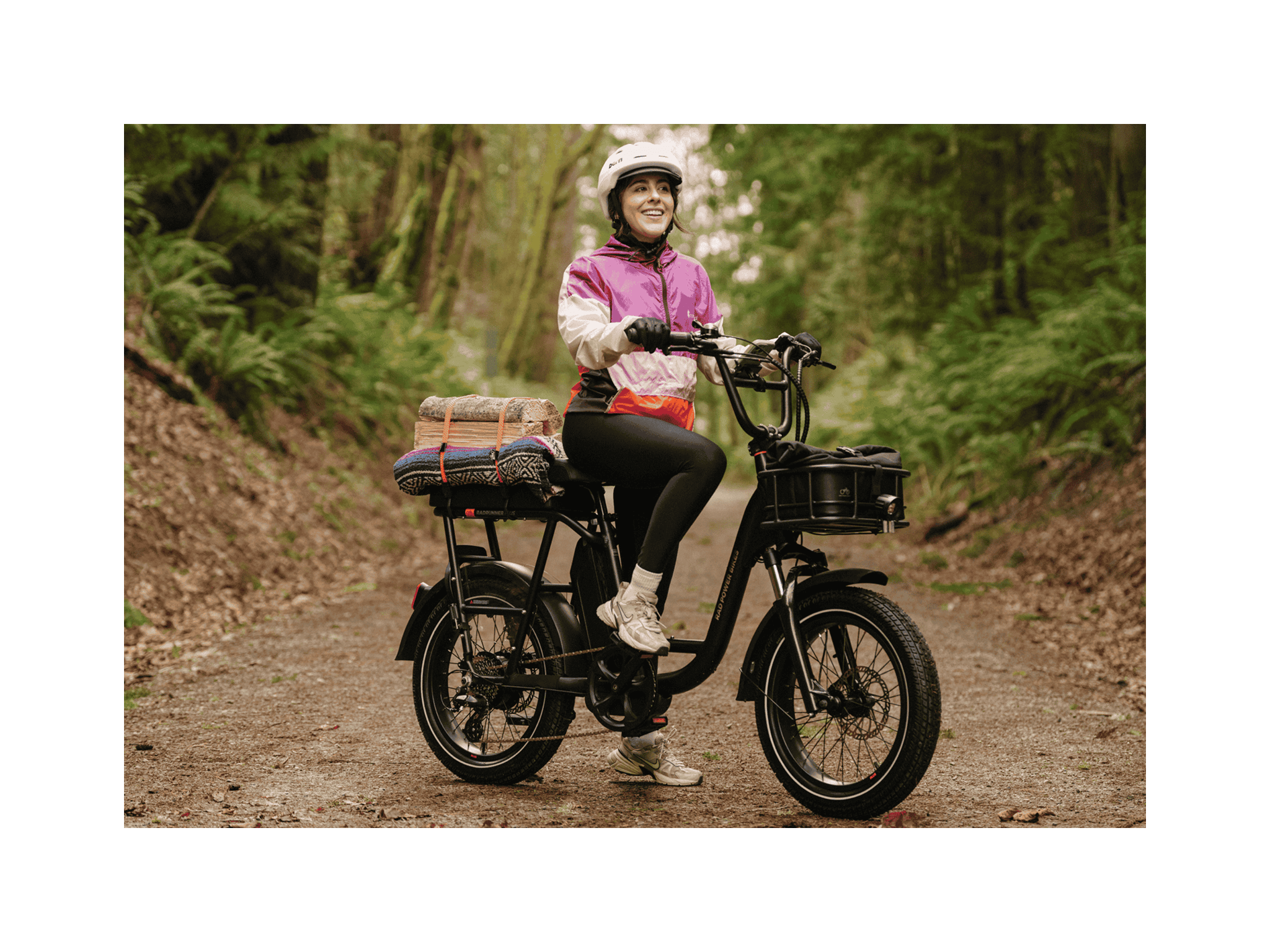 A woman sitting on a black RadRunner Plus electric cargo utility bike wearing a white helmet, pink jacket and grey pants. 