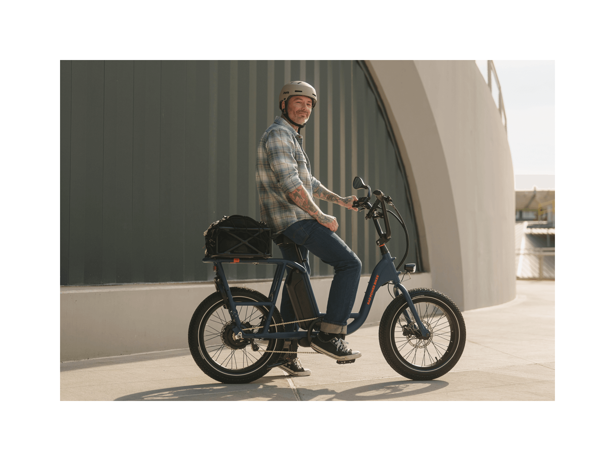 A man with tattoos wearing a plaid shirt, jeans, and a grey helmet smiling and sitting on an ebike in an urban setting. 