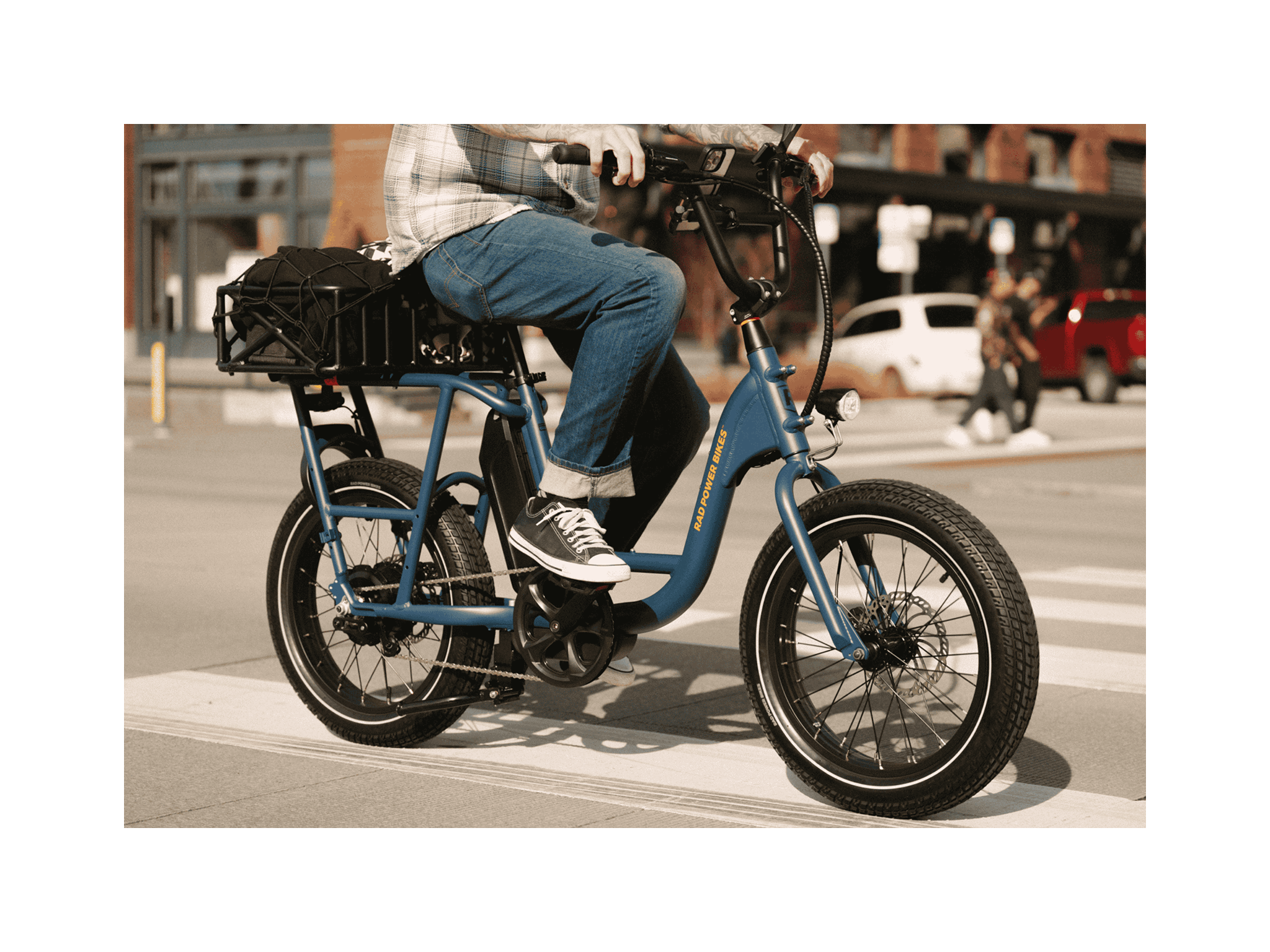A man riding a blue electric bicycle with an orange Rad Power Bikes logo, fat black tires, and large basket with a cargo net, on a paved road in an urban setting. 