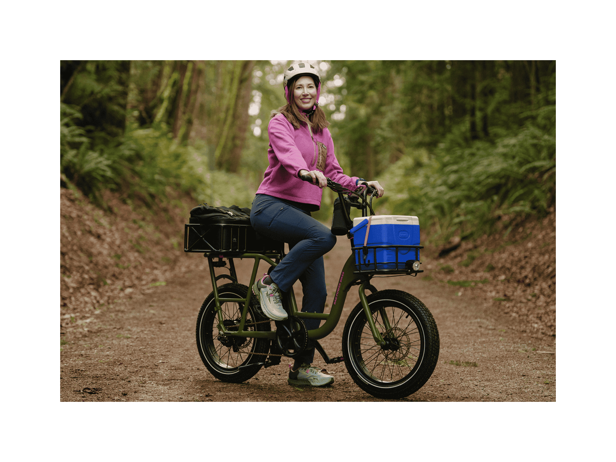 A woman on a trail in a forest sitting on a green cargo ebike with a small front basket and large rear basket wearing a pink jacket and white helmet.