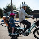 Man riding a blue RadWagon 5 electric cargo bike with two children as passengers. The children are seated with passenger accessories including the RadWagon 5 Deckpad, an orange thule yepp child seat and a caboose.