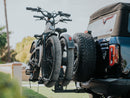 A vehicle carrying two ebikes on a rear rack drives down a road