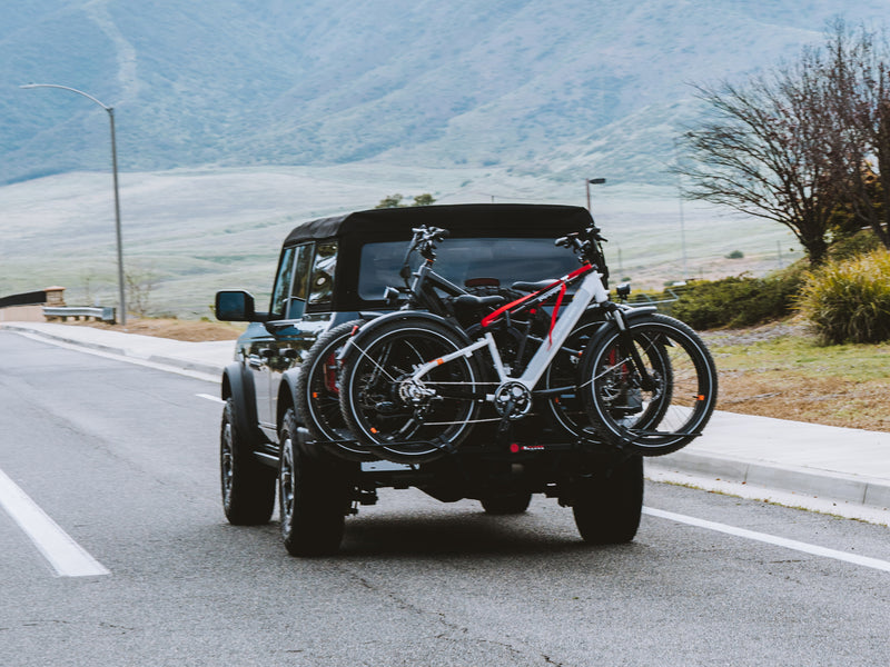 A vehicle carrying two ebikes on a rear rack drives down a road