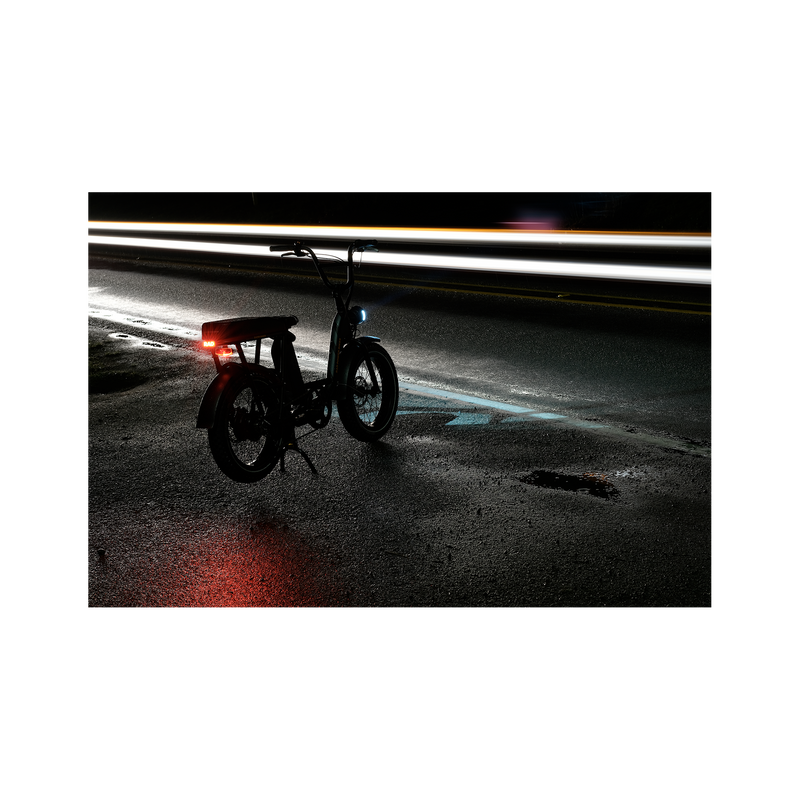 Photo of a Rad Power Bikes RadRunner electric cargo bike with passenger package parked at night on a damp street with Rad logo taillight turned on and mounted below rear seat on the rack.